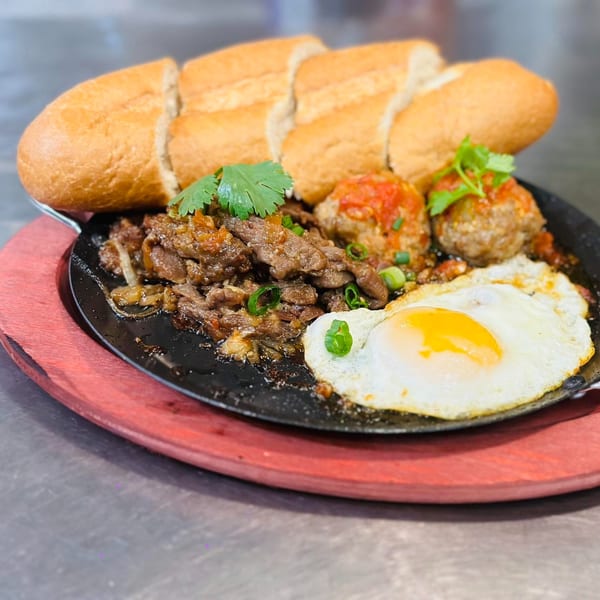 Vietnamese sizzling plate with sliced beef, meatballs, fried egg, and baguette slices on a red wooden board.