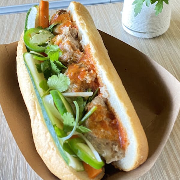 A Vietnamese banh mi sandwich with meatballs, vegetables, and sauce in a paper tray.
