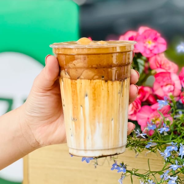 A hand holds a Vietnamese iced coffee with layered brown and white liquid, against a floral background.